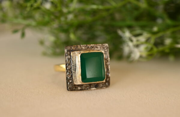The image shows a close-up of a ring. The ring has a square-shaped setting with a large, emerald-cut green stone in the center. The ring is resting on a light-colored surface, possibly a piece of fabric or paper. Soft greenery, like leaves or branches, is blurred in the background, creating a natural and elegant backdrop.
