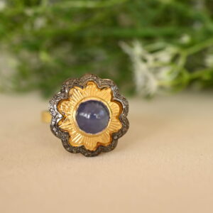 The image shows a close-up of a ring with a large, round, blue gemstone set in a gold bezel. The stone is surrounded by a halo of smaller diamonds, and the ring band is decorated with more diamonds. The background is blurred and green, possibly showing foliage. The ring appears to be resting on a light-colored surface.