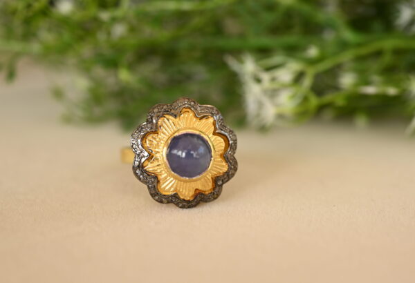 The image shows a close-up of a ring with a large, round, blue gemstone set in a gold bezel. The stone is surrounded by a halo of smaller diamonds, and the ring band is decorated with more diamonds. The background is blurred and green, possibly showing foliage. The ring appears to be resting on a light-colored surface.