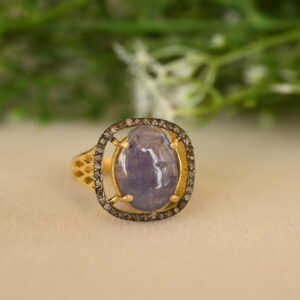 The image showcases a stunning ring featuring a large, oval-shaped blue gemstone, likely a sapphire. The sapphire is set in a delicate gold frame adorned with a halo of sparkling diamonds. The band of the ring is also crafted in gold and features a delicate, openwork design. The ring is resting on a bed of green leaves and white flowers, adding a touch of natural beauty to the scene.
