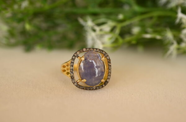The image showcases a stunning ring featuring a large, oval-shaped blue gemstone, likely a sapphire. The sapphire is set in a delicate gold frame adorned with a halo of sparkling diamonds. The band of the ring is also crafted in gold and features a delicate, openwork design. The ring is resting on a bed of green leaves and white flowers, adding a touch of natural beauty to the scene.