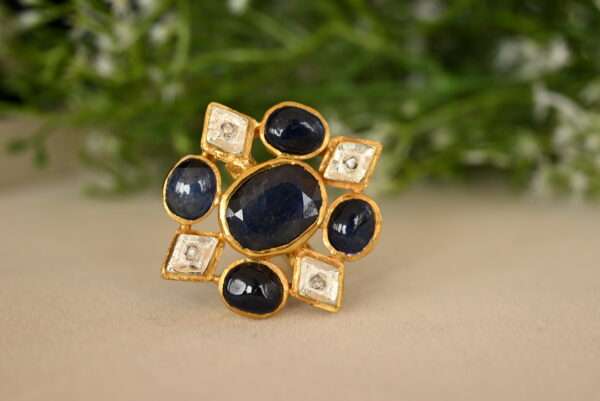 The image shows a close-up of a ring with a floral design. It appears to be made of gold and is adorned with a cluster of blue sapphires and white diamonds. The sapphires are the dominant feature, arranged in a circular pattern that resembles a flower. The diamonds are interspersed among the sapphires, adding sparkle and contrast. The ring is resting on a surface that looks like a piece of fabric with a subtle texture.