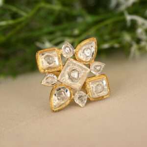 The image shows a close-up of a ring resting on a surface with a blurred background of green foliage. The ring appears to be made of silver with gold-plated accents. It features a central square-shaped design with multiple smaller diamonds set within. There are additional diamond accents around the edges of the central square. The background is blurred, suggesting a natural setting like a garden or field. Let me know if you have any other questions about the image!