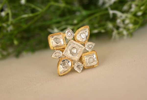 The image shows a close-up of a ring resting on a surface with a blurred background of green foliage. The ring appears to be made of silver with gold-plated accents. It features a central square-shaped design with multiple smaller diamonds set within. There are additional diamond accents around the edges of the central square. The background is blurred, suggesting a natural setting like a garden or field. Let me know if you have any other questions about the image!