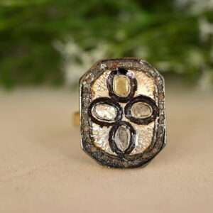 The ring has a rectangular or octagonal shape. It is adorned with multiple small diamonds, possibly set in silver. The background appears to be a light-colored surface, possibly a fabric or table. There are green leaves or branches visible in the background, suggesting a natural setting.