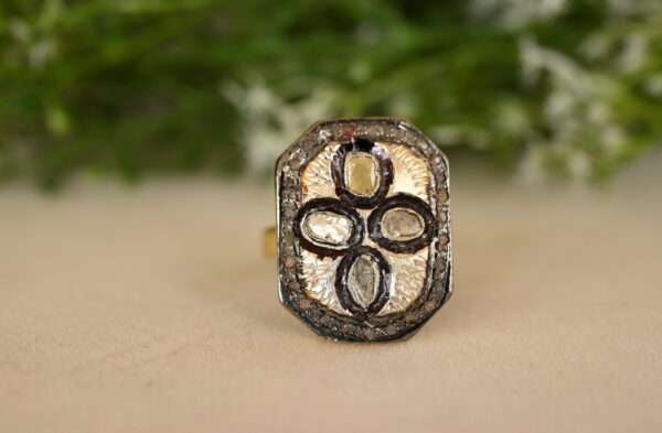 The ring has a rectangular or octagonal shape. It is adorned with multiple small diamonds, possibly set in silver. The background appears to be a light-colored surface, possibly a fabric or table. There are green leaves or branches visible in the background, suggesting a natural setting.