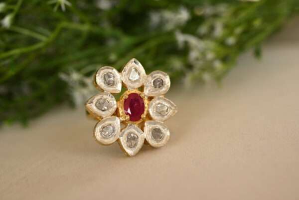 This image features a stunning ring set in yellow gold, adorned with a vibrant red ruby as its centerpiece. The ruby is surrounded by a halo of shimmering, rose-cut polki diamonds, which are traditionally uncut and have a soft, matte finish. The ring's design is reminiscent of vintage Indian jewelry, with its intricate detailing and delicate craftsmanship.