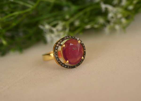 The image shows a close-up of a beautiful ring. It features a large, oval-shaped ruby gemstone set in a gold-colored setting. The ruby is a deep, rich red color and appears to be of high quality. Surrounding the ruby is a halo of smaller, sparkling diamonds. The band of the ring appears to be made of gold or a gold-plated metal. The background of the image is a soft, blurred green and white, possibly flowers or foliage.