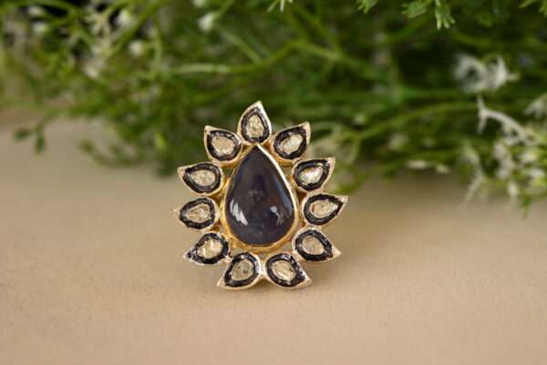 The image shows a ring with a large, pear-shaped blue gemstone, likely a sapphire, surrounded by a halo of smaller diamonds. The ring appears to be made of silver or gold and has intricate floral designs. It's a beautiful and elegant piece of jewelry.