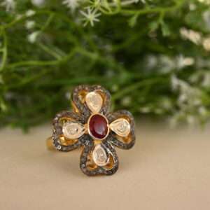 The image showcases a stunning ring adorned with a vibrant red ruby as the centerpiece. The ruby is surrounded by a halo of sparkling white diamonds, creating a dazzling contrast. The ring appears to be made of silver or gold, and the intricate design suggests it might be inspired by vintage or antique styles. The background of the image features lush green foliage with delicate white flowers, adding a touch of nature to the overall aesthetic.