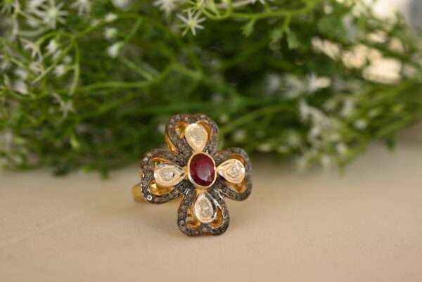 The image showcases a stunning ring adorned with a vibrant red ruby as the centerpiece. The ruby is surrounded by a halo of sparkling white diamonds, creating a dazzling contrast. The ring appears to be made of silver or gold, and the intricate design suggests it might be inspired by vintage or antique styles. The background of the image features lush green foliage with delicate white flowers, adding a touch of nature to the overall aesthetic.