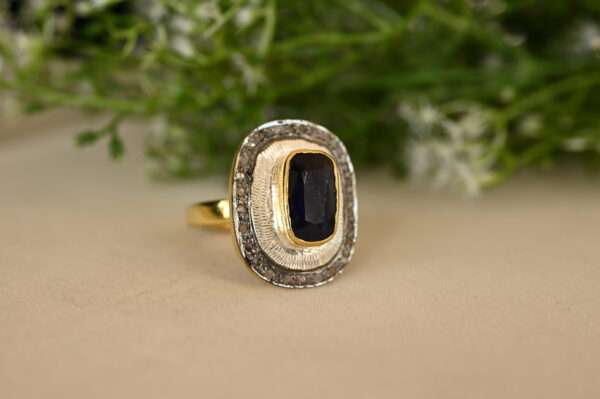The image shows a ring with a large, rectangular sapphire gemstone set in a gold and silver band. The sapphire is a deep blue color and appears to be faceted. The band is a mix of yellow gold and oxidized silver, creating a unique and contrasting look. The ring is resting on a beige surface with some greenery in the background.