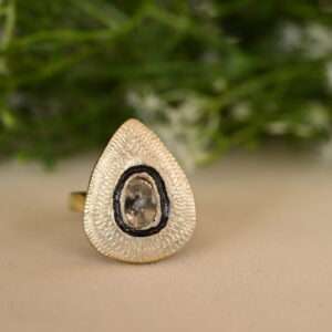 The image shows a close-up of a ring with a teardrop-shaped setting. The setting is made of gold or a gold-toned metal and has intricate, textured details. Inside the setting is a clear gemstone, possibly a diamond or a crystal. The background is blurred and appears to be a cluster of small white flowers and green leaves.