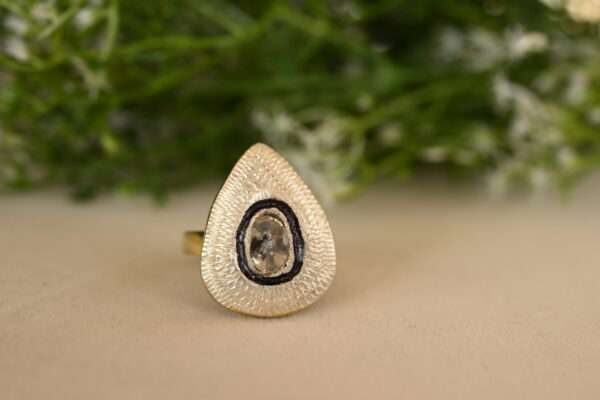 The image shows a close-up of a ring with a teardrop-shaped setting. The setting is made of gold or a gold-toned metal and has intricate, textured details. Inside the setting is a clear gemstone, possibly a diamond or a crystal. The background is blurred and appears to be a cluster of small white flowers and green leaves.