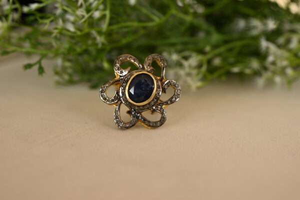 The image shows a close-up of a beautiful ring resting on a textured surface. The ring is made of silver with a dark oxidized finish, creating a striking contrast. It features a large, oval-shaped blue sapphire as the centerpiece. The sapphire is surrounded by a delicate halo of smaller diamonds, which sparkle and catch the light. The ring's band is adorned with intricate floral motifs, adding to its overall elegance. The background is blurred and features a soft green color, possibly from foliage, which adds a natural touch to the image.