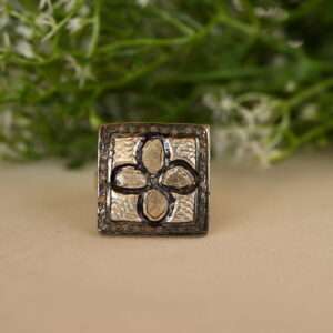 The image shows a silver ring resting on a beige surface. The ring has a square-shaped center with a floral pattern. Inside the square, four diamond-shaped petals are visible, each set with a diamond. The background is blurred and features green foliage with small white flowers.