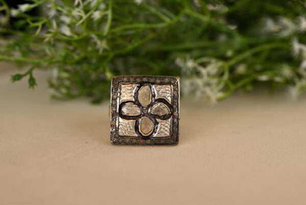 The image shows a silver ring resting on a beige surface. The ring has a square-shaped center with a floral pattern. Inside the square, four diamond-shaped petals are visible, each set with a diamond. The background is blurred and features green foliage with small white flowers.