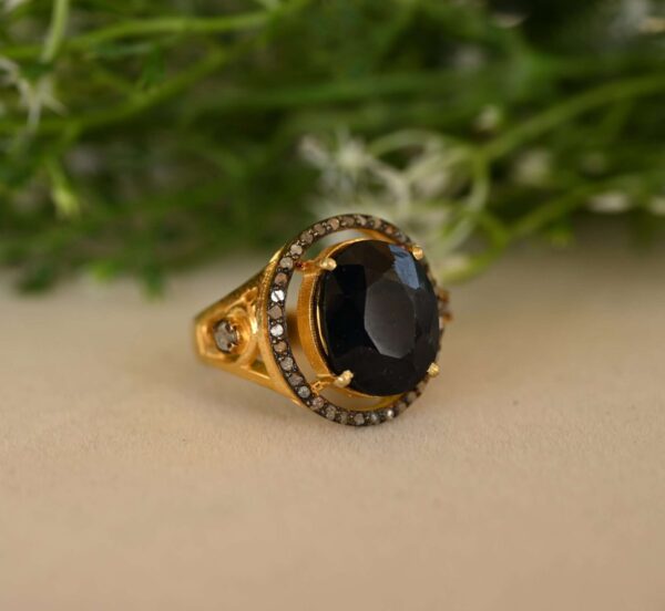 The image shows a close-up of a beautiful ring resting on a surface. Ring: The ring appears to be made of gold with a large, oval-shaped black stone in the center. The stone is likely onyx. Surrounding the onyx is a halo of smaller, sparkling white stones, most likely diamonds. Background: The background is blurred, but it seems to be a natural setting with some greenery visible.