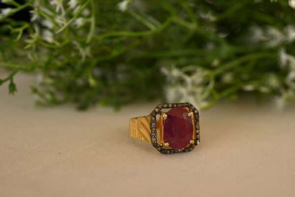 The image shows a close-up of a ring resting on a beige surface, likely a table or counter. The ring has a prominent oval-shaped ruby gemstone in the center, surrounded by a halo of smaller black diamonds. The ring's band is made of gold and has a textured pattern. The background is blurred and features a sprig of green foliage with small white flowers.