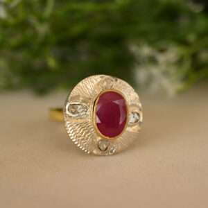 The image shows a close-up of a ring. It appears to be made of yellow gold and features a large, oval-shaped ruby as the centerpiece. The ruby is a deep, rich red color and is surrounded by a halo of smaller diamonds. The ring has a delicate, flowing design, with the band tapering towards the ruby.