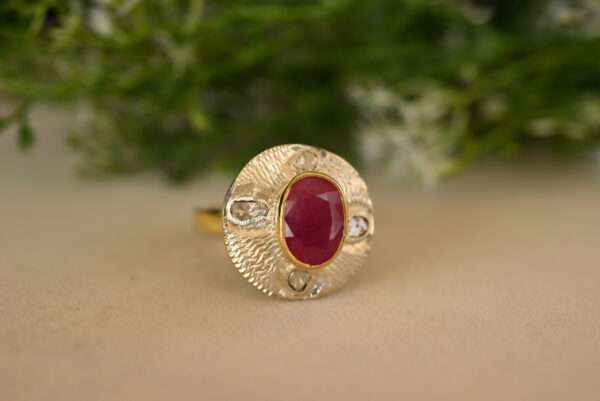 The image shows a close-up of a ring. It appears to be made of yellow gold and features a large, oval-shaped ruby as the centerpiece. The ruby is a deep, rich red color and is surrounded by a halo of smaller diamonds. The ring has a delicate, flowing design, with the band tapering towards the ruby.