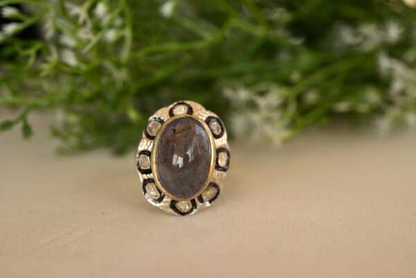 The image shows a close-up of a ring resting on a textured surface. The ring appears to be made of silver and features a large oval-shaped gemstone in the center. The gemstone is a light to medium shade of gray with hints of blue and purple, possibly a sapphire or a smoky quartz. "Moonstone & Diamond Ring"