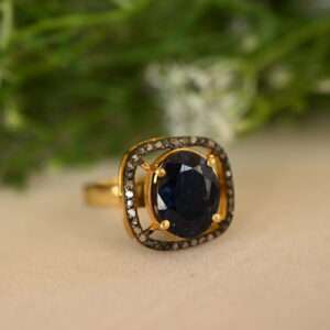 The image shows a close-up of a beautiful ring. The ring appears to be made of gold and features a large, oval-shaped blue sapphire as the centerpiece. The sapphire is surrounded by a halo of smaller diamonds, which add sparkle and enhance the brilliance of the main stone. The ring is resting on a surface that looks like a piece of fabric, possibly velvet or satin. The background is blurred and appears to be green, suggesting the presence of foliage or plants.