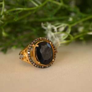 The image shows a close-up of a ring with a large, oval-shaped black sapphire gemstone. The sapphire is set in a gold-colored setting, which is embellished with smaller diamonds. The ring is resting on a bed of green leaves and white flowers.