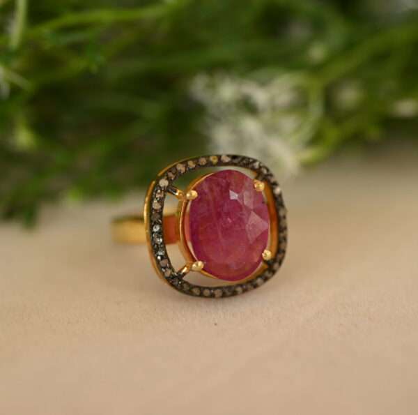 The image shows a close-up of a ring with a large, oval-shaped ruby gemstone set in a gold band. The ruby is a deep, rich red color and is surrounded by a halo of smaller, sparkling diamonds. The band of the ring is also decorated with additional diamonds. The background of the image is blurred and appears to be green foliage.