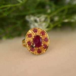 The image shows a close-up of a gold ring with a large, oval-shaped ruby in the center. The ruby is a deep, rich red color and is surrounded by a halo of smaller, round rubies. The ring band is also gold and appears to be intricately designed with small, decorative details. The background of the image is blurred and appears to be a bed of green leaves and white flowers, suggesting a natural setting.
