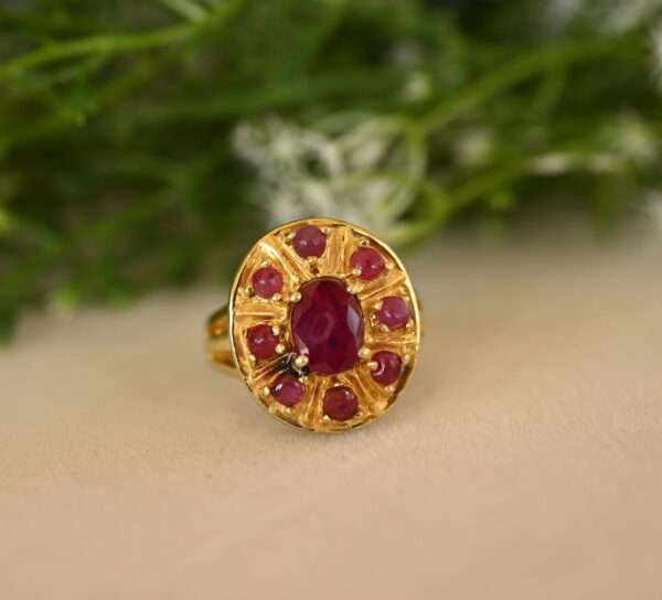 The image shows a close-up of a gold ring with a large, oval-shaped ruby in the center. The ruby is a deep, rich red color and is surrounded by a halo of smaller, round rubies. The ring band is also gold and appears to be intricately designed with small, decorative details. The background of the image is blurred and appears to be a bed of green leaves and white flowers, suggesting a natural setting.