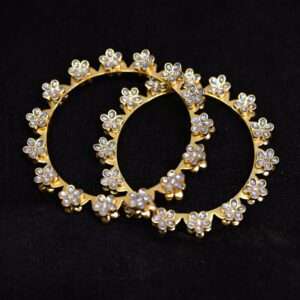 The image shows a pair of gold-toned bangles adorned with a delicate floral motif. Each flower is meticulously crafted with small, sparkling stones, likely cubic zirconia or pearls. The bangles are resting on a dark background.