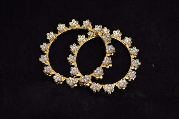 The image shows a pair of gold-toned bangles adorned with a delicate floral motif. Each flower is meticulously crafted with small, sparkling stones, likely cubic zirconia or pearls. The bangles are resting on a dark background.