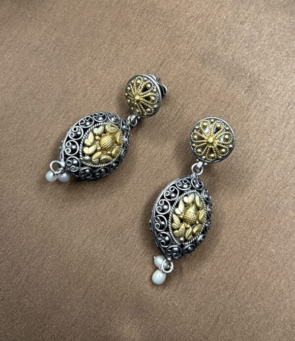 "A close-up of a pair of black and gold fusion earrings. The earrings have a round, ornate design with a central gold motif. Delicate pearls hang from the bottom of each earring."