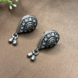 The image shows a pair of silver earrings with a teardrop shape. The earrings have an oxidized finish, giving them an antique look. The teardrop is adorned with intricate floral patterns and has two small dangling beads at the bottom. The earrings are resting on a brown surface with a small green plant in the background.