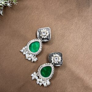 The image shows a pair of elegant fusion earrings. They feature a square-shaped top adorned with intricate silver filigree work and embellished with sparkling white stones. From the center of the square, a delicate pear-shaped green gemstone dangles gracefully. Below the gemstone, a cluster of lustrous white pearls adds a touch of elegance and sophistication. The earrings are resting on a brown surface, possibly a table or a piece of cloth.