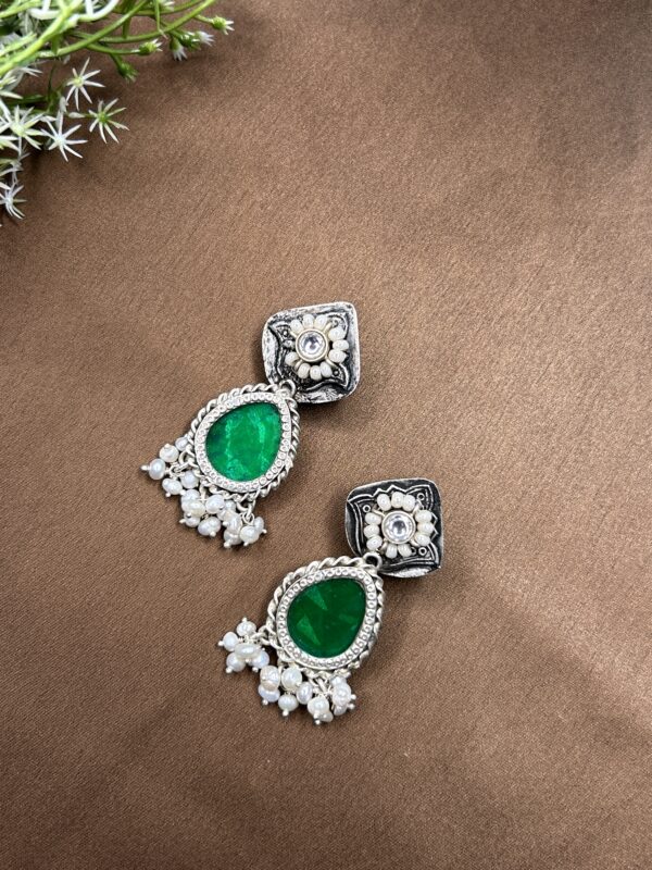 The image shows a pair of elegant fusion earrings. They feature a square-shaped top adorned with intricate silver filigree work and embellished with sparkling white stones. From the center of the square, a delicate pear-shaped green gemstone dangles gracefully. Below the gemstone, a cluster of lustrous white pearls adds a touch of elegance and sophistication. The earrings are resting on a brown surface, possibly a table or a piece of cloth.