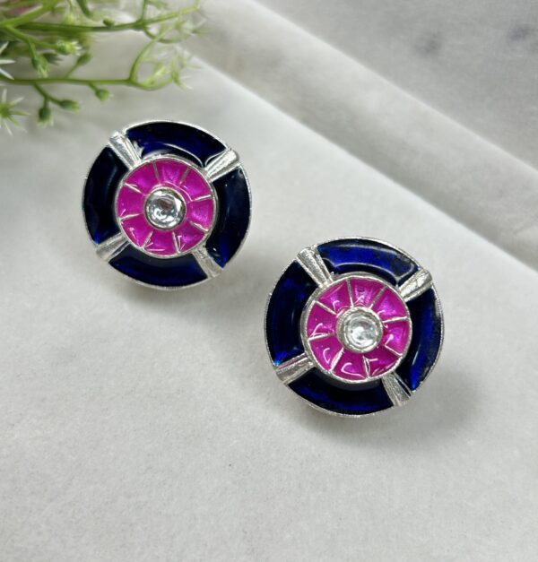 The image shows a pair of oval-shaped stud earrings with a unique design. Colors: The earrings feature a striking color combination of deep blue, vibrant pink, and white. Design: The central part of each earring has a white stone, surrounded by a circular pink enamel inlay. This pink circle is then framed by a wider band of deep blue enamel. Material: The earrings appear to be made of silver metal.