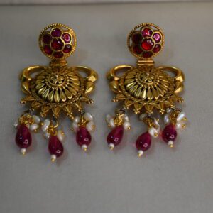 The image shows a pair of gold earrings with a traditional Indian design. The earrings are made of gold and feature a circular motif adorned with intricately set red stones. Below the circular motif, there is a cluster of pearls and red stones hanging down, creating a beautiful and elegant drop. The earrings are likely handmade and showcase intricate goldwork.