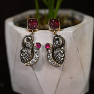 The image shows a pair of elegant earrings. The earrings feature a striking peacock motif, a symbol of grace and beauty in Indian culture. The peacock is intricately carved and adorned with sparkling white stones, adding a touch of elegance. The earrings are further embellished with a vibrant red stone at the top, creating a stunning contrast. Earring