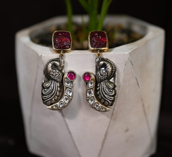 The image shows a pair of elegant earrings. The earrings feature a striking peacock motif, a symbol of grace and beauty in Indian culture. The peacock is intricately carved and adorned with sparkling white stones, adding a touch of elegance. The earrings are further embellished with a vibrant red stone at the top, creating a stunning contrast. Earring