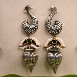 The image shows a pair of elegant earrings with a peacock motif. The earrings are made with silver and feature intricate detailing on the peacock's feathers and head. The peacock's head is adorned with a small green gemstone, adding a touch of color. The earrings also have a dangling element with small pearls and a green gemstone accent.