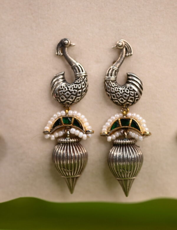 The image shows a pair of elegant earrings with a peacock motif. The earrings are made with silver and feature intricate detailing on the peacock's feathers and head. The peacock's head is adorned with a small green gemstone, adding a touch of color. The earrings also have a dangling element with small pearls and a green gemstone accent.