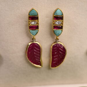The image shows a pair of elegant earrings. The earrings have a gold base and feature a combination of colorful gemstones, including turquoise, ruby, and diamonds. The earrings also have a delicate leaf motif, symbolizing growth and nature. The overall design of the earrings is a fusion of traditional Indian craftsmanship and contemporary style.