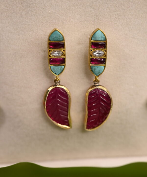 The image shows a pair of elegant earrings. The earrings have a gold base and feature a combination of colorful gemstones, including turquoise, ruby, and diamonds. The earrings also have a delicate leaf motif, symbolizing growth and nature. The overall design of the earrings is a fusion of traditional Indian craftsmanship and contemporary style.