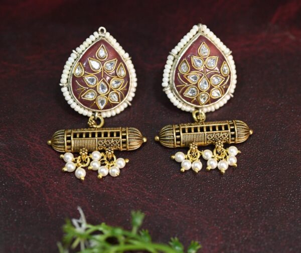 The image showcases a pair of elegant fusion earrings. They feature a teardrop-shaped pendant adorned with a beautiful floral motif in a rich, warm shade of purple. The intricate detailing of the flowers is truly captivating, with each petal meticulously crafted and embellished with sparkling white stones.