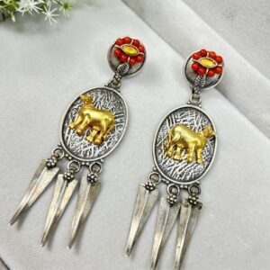 The image shows a pair of traditional ethnic-style earrings. They feature an oval silver base with a golden cow design in the center. The top part has red bead embellishments around a central yellow gem, and three pointed silver dangles hang from each earring, giving them a tribal look.