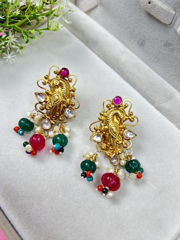This image shows a pair of ornate, traditional Indian earrings. The earrings are gold-toned with intricate peacock designs, embedded with white stones and topped with a round pink gem. They are adorned with hanging green and pink beads, tiny pearls, and multicolored accent beads, giving them a festive and vibrant look.