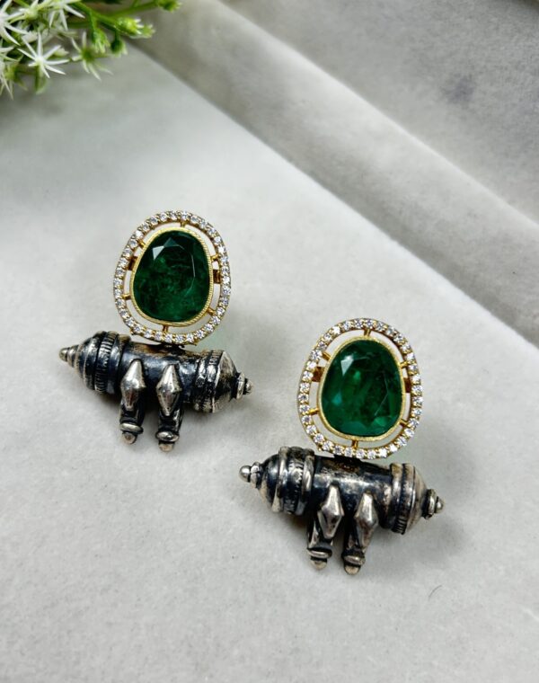 The image showcases a pair of elegant earrings. The earrings feature a striking green gemstone, likely an emerald, set in a delicate gold frame. The frame is adorned with sparkling white stones, adding a touch of glamour. The lower part of the earrings incorporates a silver-toned element with intricate detailing, reminiscent of traditional Indian jewelry motifs. The combination of the green gemstone, gold frame, and silver accents creates a truly unique and eye-catching look.