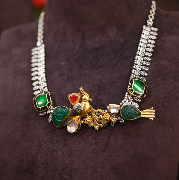 The image appears to be a close-up shot of a necklace resting on a dark, textured surface. The necklace has a distinct ethnic or tribal feel with its use of colorful stones and intricate metalwork. Necklace: The necklace is composed of a mix of materials. There are several large, vibrant green stones, likely gemstones or glass, set in what looks like silver or white metal. The metalwork is detailed, with intricate patterns and designs visible. A small, gold-colored bird motif is incorporated into the design. Background: The necklace is placed on a dark surface with a textured appearance, possibly velvet or a similar fabric. This provides a good contrast to the colors of the necklace.